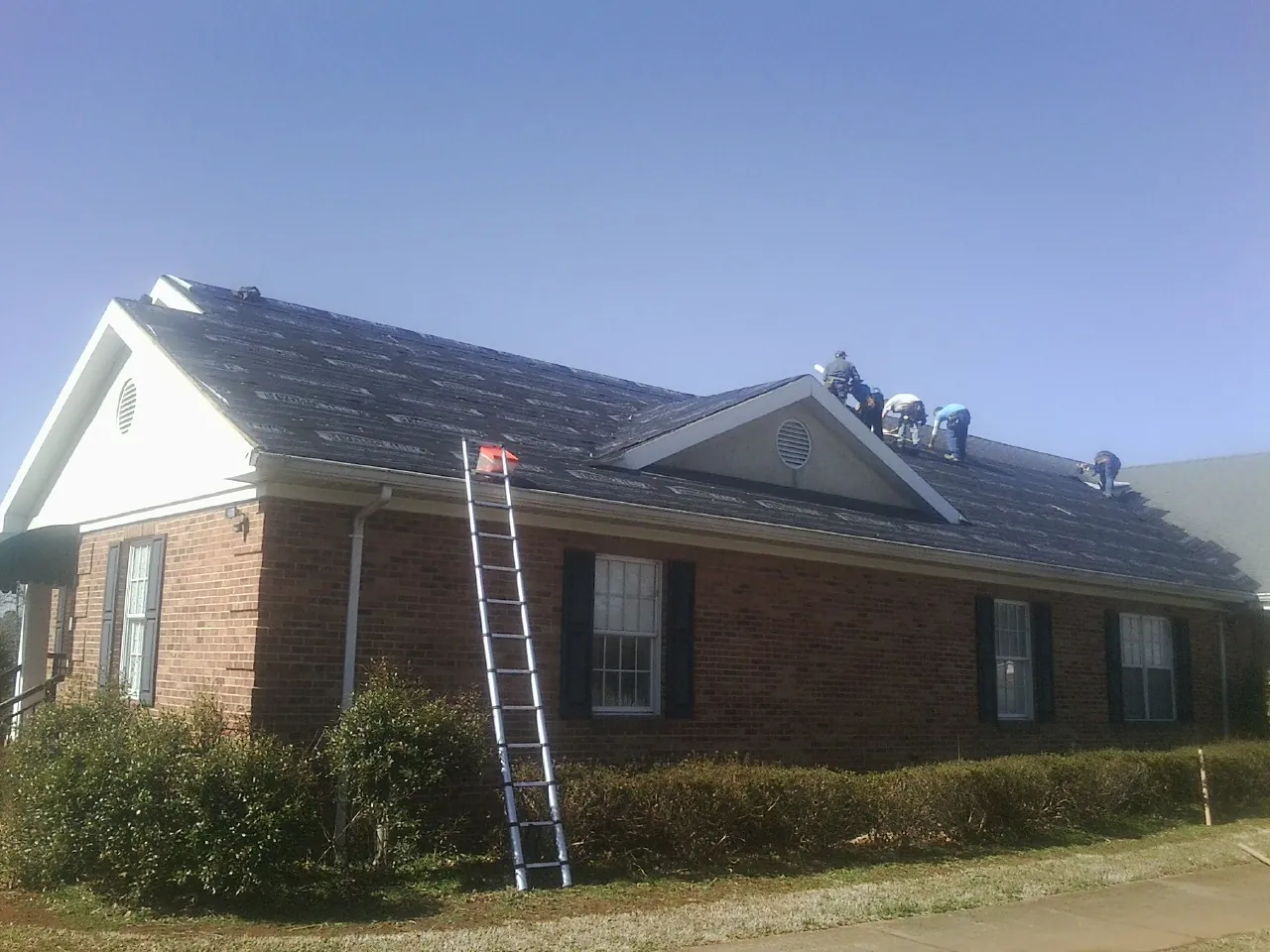 Crew tearing shingles off a brick ranch roof with a ladder set — Metal Roof Repair work by our crew in Manton