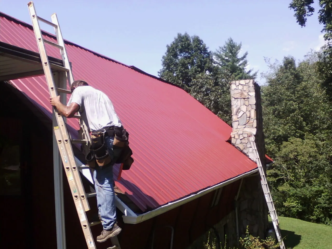 Roofer on a ladder beside a red metal roof and stone chimney — Soffit & Fascia Repair by Roofing Brothers in Manton